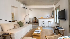 A modern living room with a sofa, coffee table, and a fireplace. There is a kitchen area with a bar and stools. - The River House (Ula)