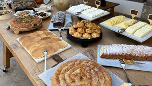An outdoor buffet with various food items, including pastries, cheeses, and fruits, set up on a wooden table under stone pillars. - The River House (Ula)