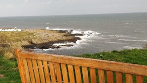 Um banco de madeira na relva com vista para um litoral rochoso, com ondas a quebrar nas rochas.