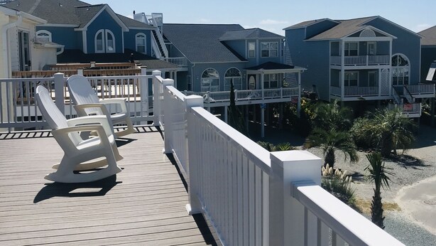 Una terraza de madera con barandas blancas y sillas de lounge, con vista a una propiedad frente a la playa con casas azules y un cielo despejado.
