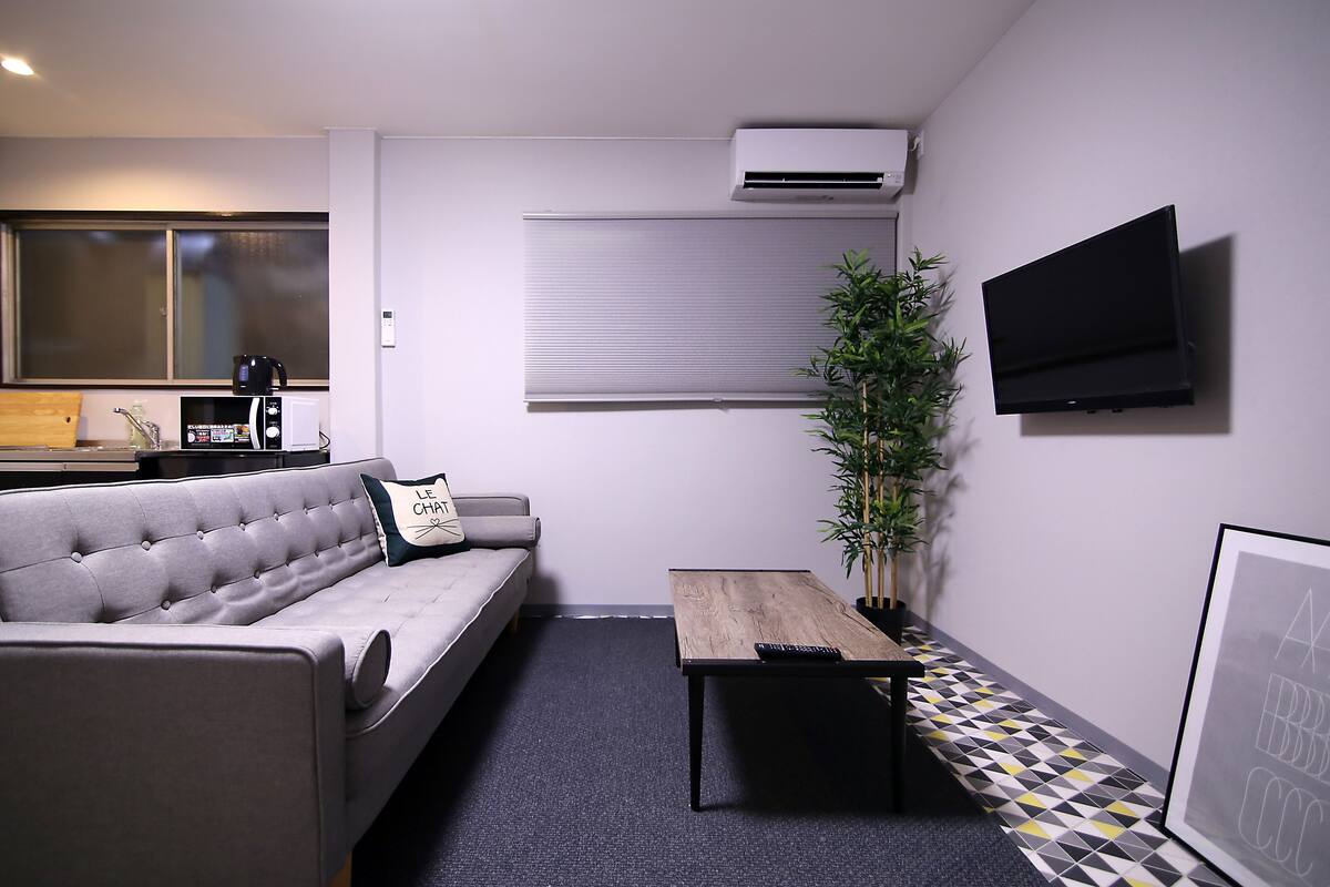 A modern living room with a grey sofa, a wooden coffee table, a potted plant, and a mounted TV.