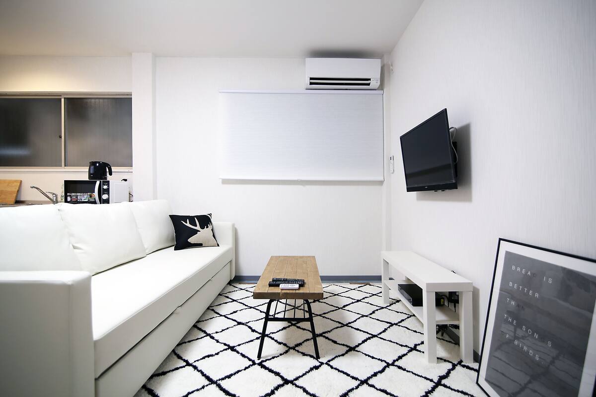 A modern living room with a white couch, a coffee table, a flat-screen TV mounted on the wall, and a window with blinds.