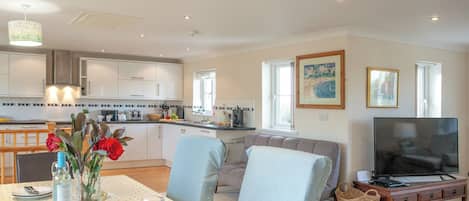 A modern kitchen with white cabinets, a dining area with a table set for two, and a living room with a sofa and a TV.