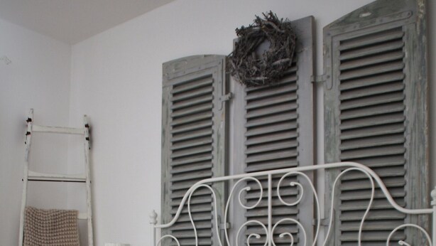 A bed with a white metal frame, grey patterned pillows, and a white throw blanket. There is a small bedside table with a lamp and a vase of flowers. A ladder is leaning against the wall, and a wreath hangs above the bed.