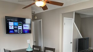 A dining area with a wooden table and chairs, a ceiling fan, a flat-screen TV mounted on the wall, and a staircase leading to another room. - Relax close to the lakefront! Three bedroom Avon Lake retreat just steps away. (AVON LAKE)