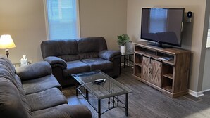 A living room with a dark brown leather sofa, a glass coffee table, a wooden entertainment unit with a flat-screen TV, and a ceiling fan. - Relax close to the lakefront! Three bedroom Avon Lake retreat just steps away. (AVON LAKE)