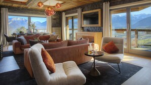 A spacious living room with a large sofa, armchairs, and a dining area. A fireplace is visible, and the room has large windows offering mountain views.