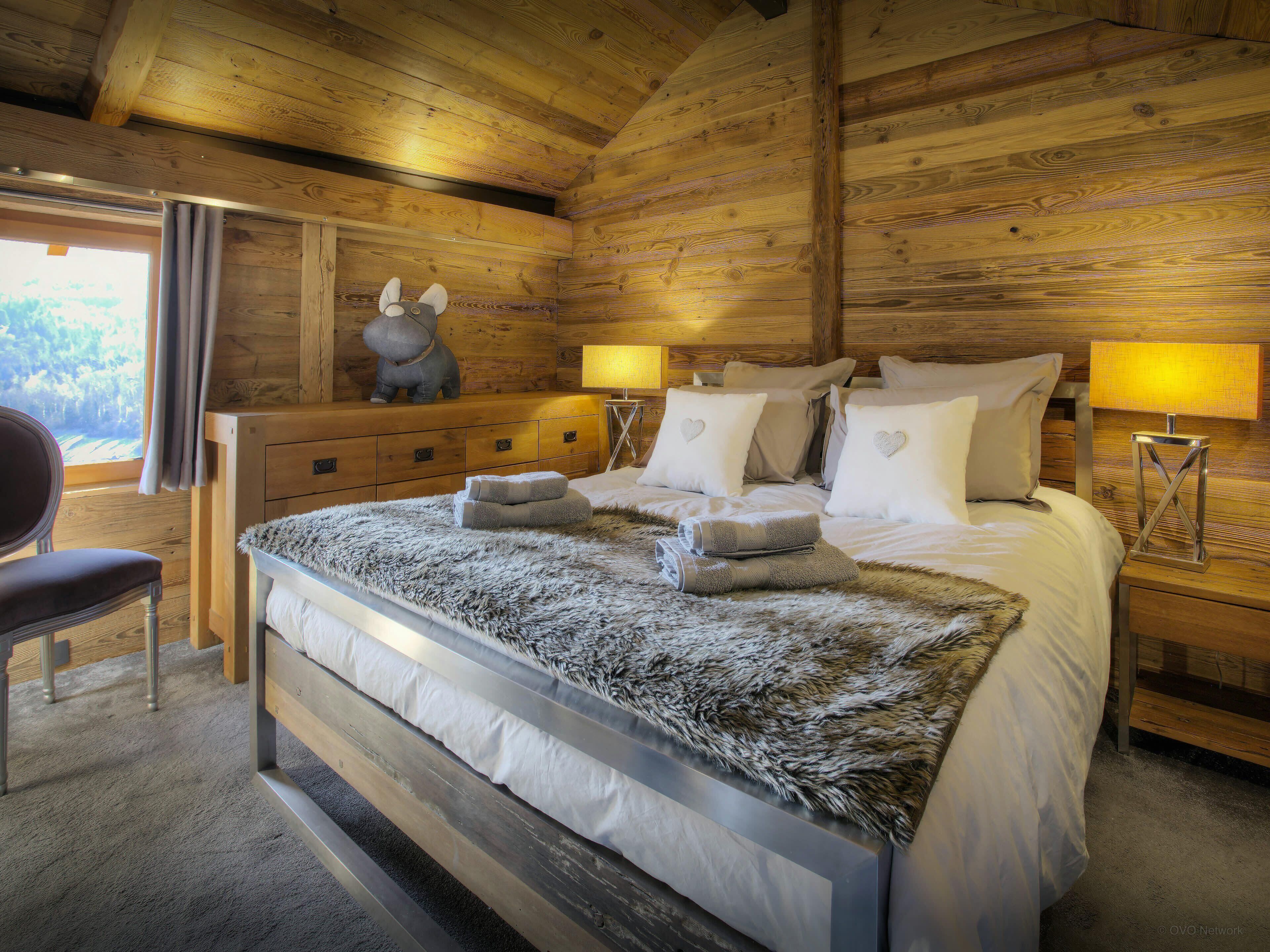 A wooden cabin bedroom with a bed, two bedside lamps, a chair, and a dresser.