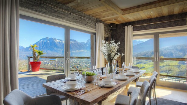 A dining area with a wooden table set for six, large windows offering mountain views, and a potted plant on the balcony.