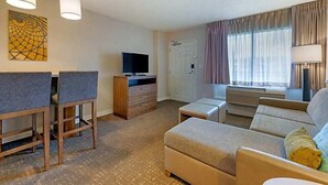Une chambre d’hôtel équipée d’un canapé, d’une télévision, d’un bureau avec deux chaises, d’une petite table et d’une fenêtre avec des rideaux.
