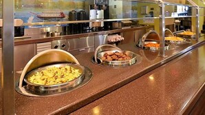 Un buffet proposant divers plats présentés dans des plats de service en métal, une machine à café et une vue sur un bateau à travers une fenêtre en verre.