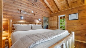 A wooden cabin bedroom with a bed, wooden walls, a door, and a window. - Cute, Cozy Log Cabin, Private setting, Decorated in red & black Rooster theme. (Rogers)