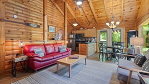 A cozy cabin interior with a red leather sofa, wooden dining table, and a kitchen area. - Cute, Cozy Log Cabin, Private setting, Decorated in red & black Rooster theme. (Rogers)