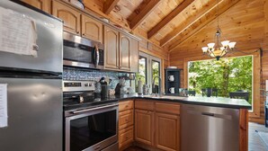 A kitchen with wooden cabinets, stainless steel appliances, and a view of greenery outside. - Cute, Cozy Log Cabin, Private setting, Decorated in red & black Rooster theme. (Rogers)