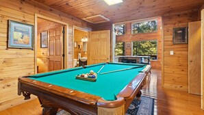 Een houten pooltafel met ballen, een poolkeu en een vloerkleed in een ruimte met houten wanden en grote ramen.