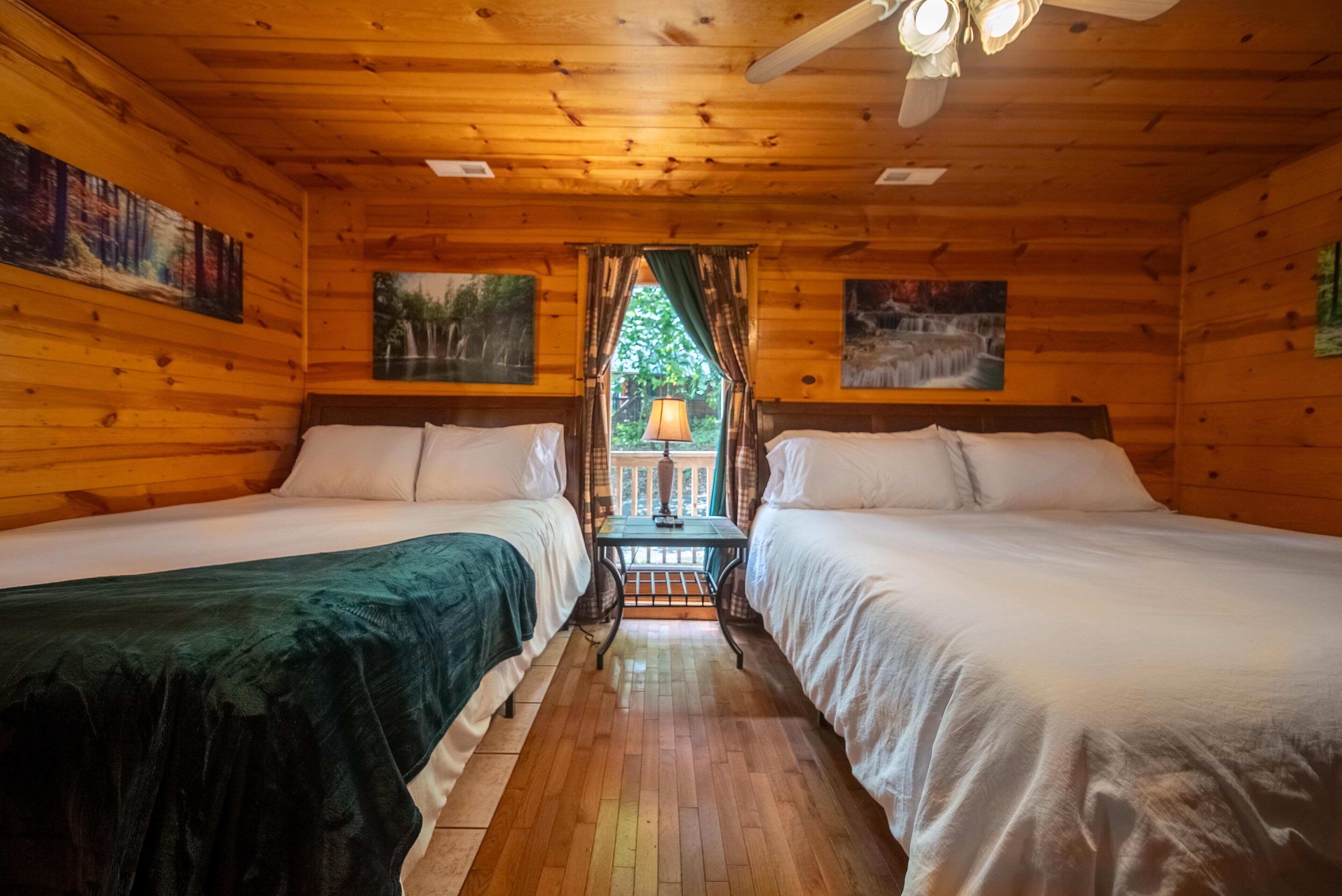 A cabin with two beds, wooden walls, a window with curtains, and a table with a lamp.