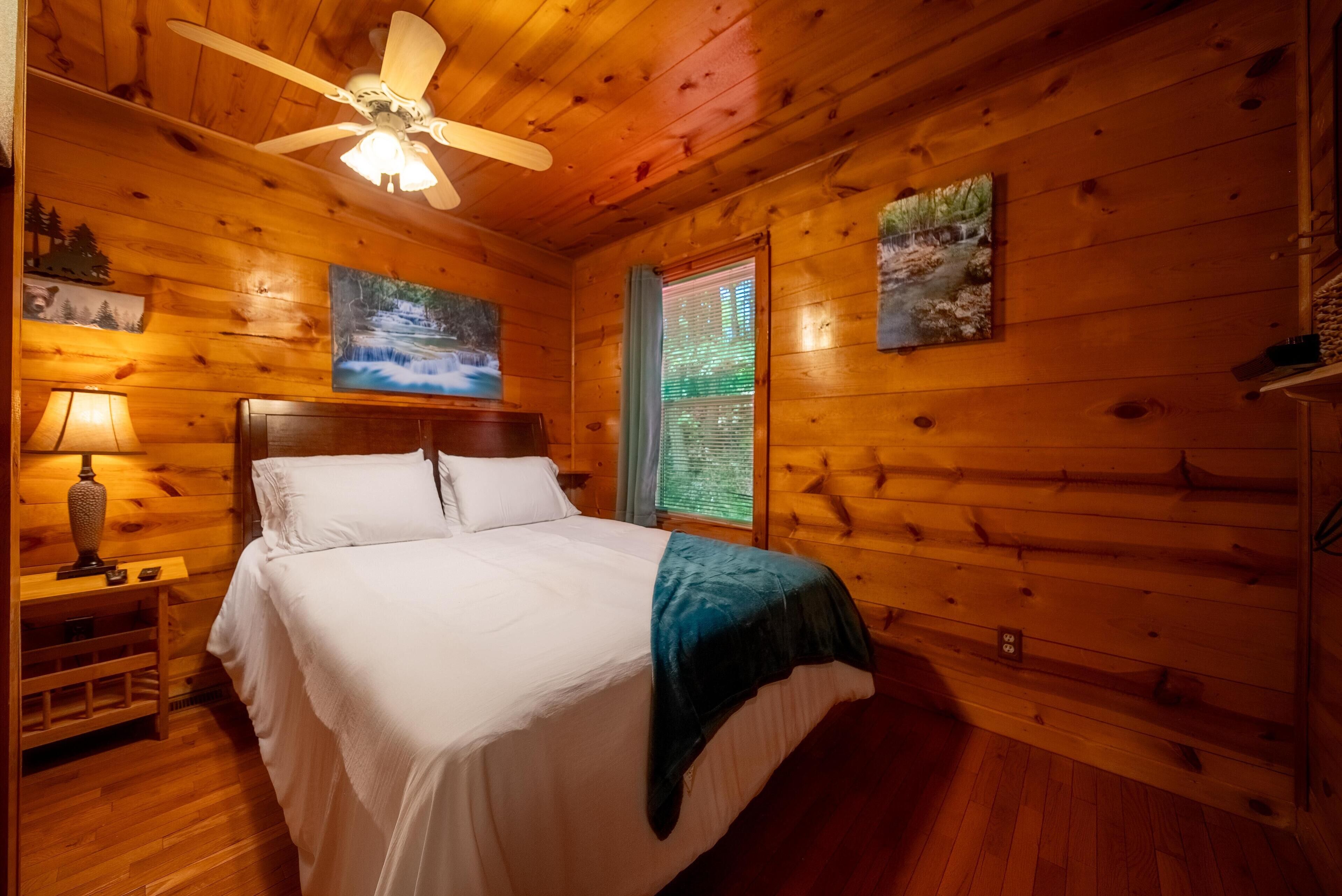 A wooden cabin bedroom with a bed, a nightstand, a lamp, a ceiling fan, and two paintings on the walls.