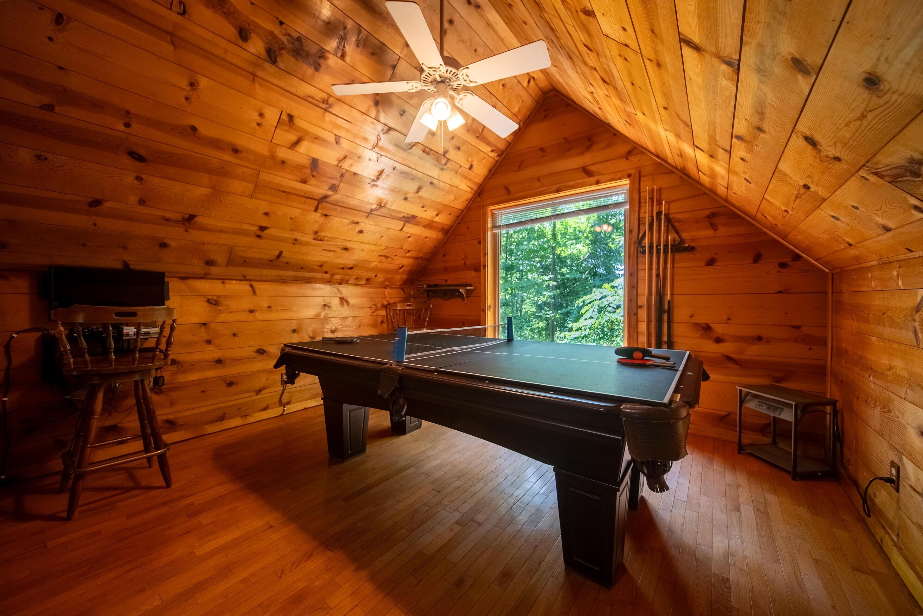 Een houten kamer met een pooltafel, een plafondventilator en een raam met uitzicht op bomen.