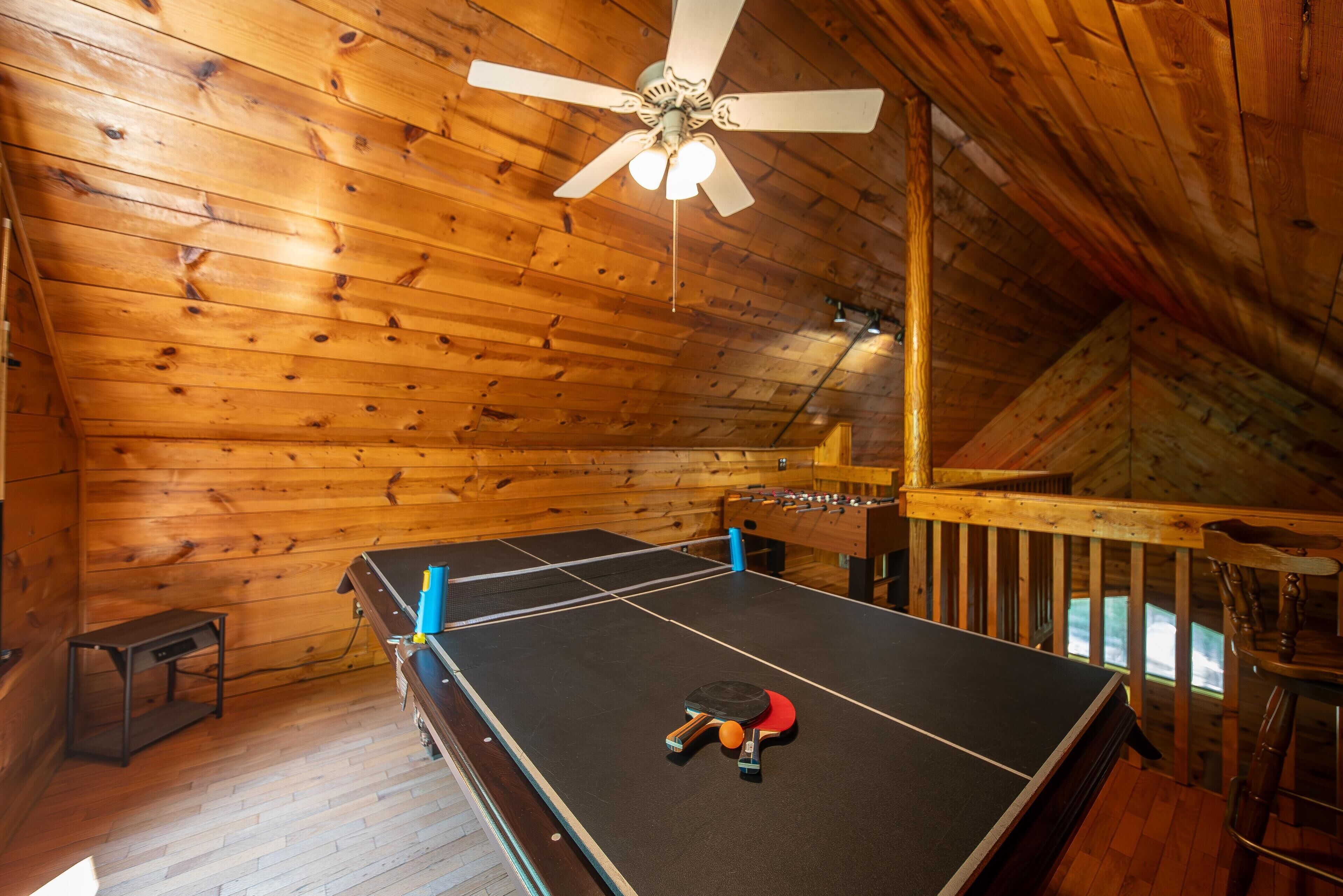 Una habitación de madera con mesa de ping pong, ventilador de techo y escalera de madera.