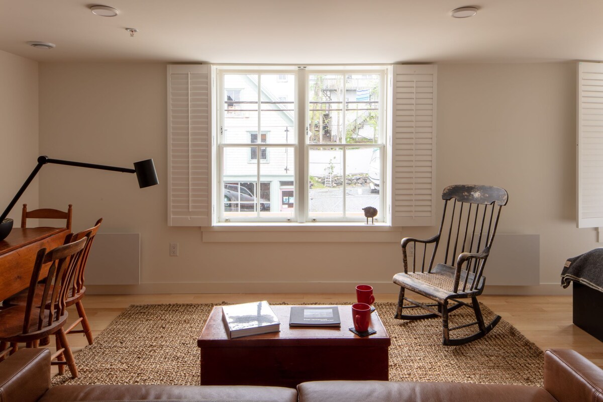 Ein Wohnzimmer mit einem Holzesstisch, einem Schaukelstuhl, einem Couchtisch mit Büchern und Tassen sowie einem großen Fenster mit Jalousien.