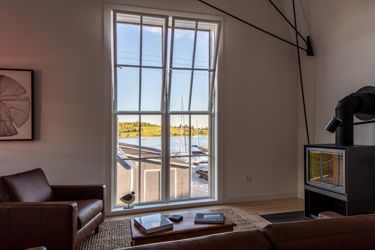 Ein Wohnzimmer mit großem Fenster, einem braunen Ledersofa, einem Holztisch und einem schwarzen Kamin.