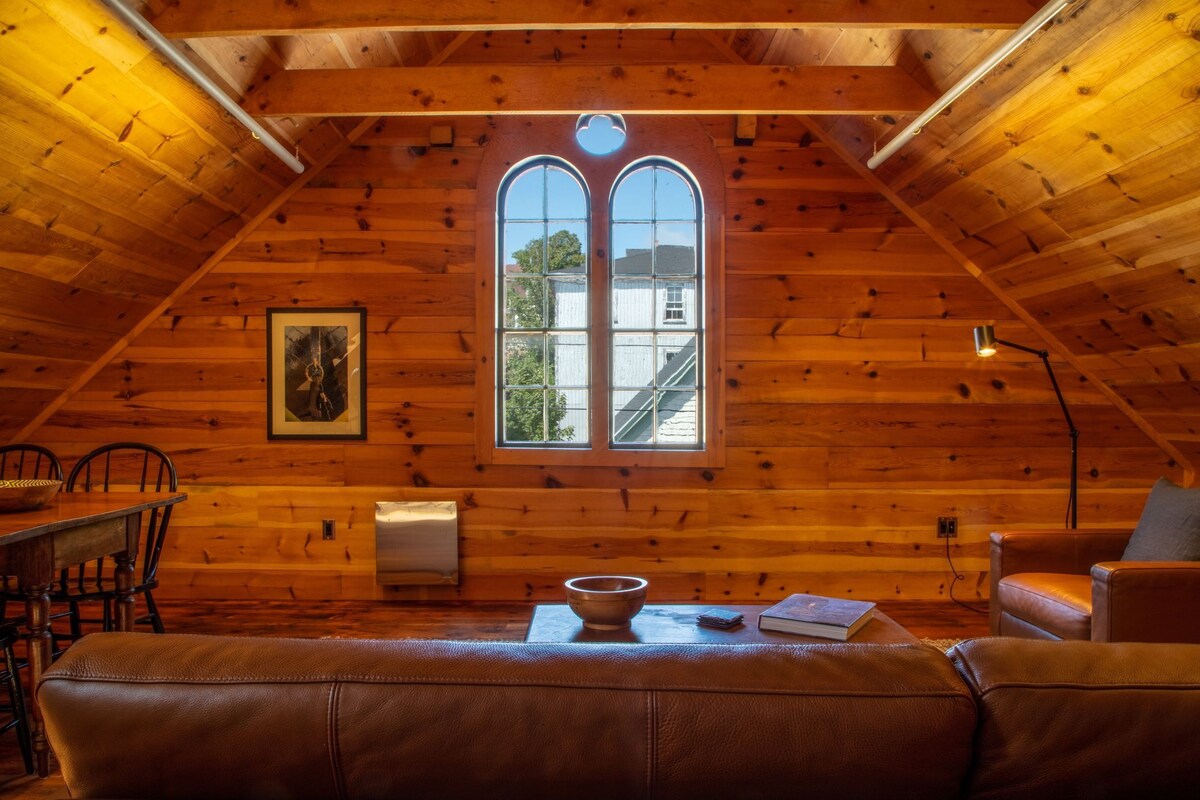 Ein gemütliches Dachzimmer mit Holzwänden, einem großen Bogenfenster, einem Esstisch und Ledersitzmöbeln.