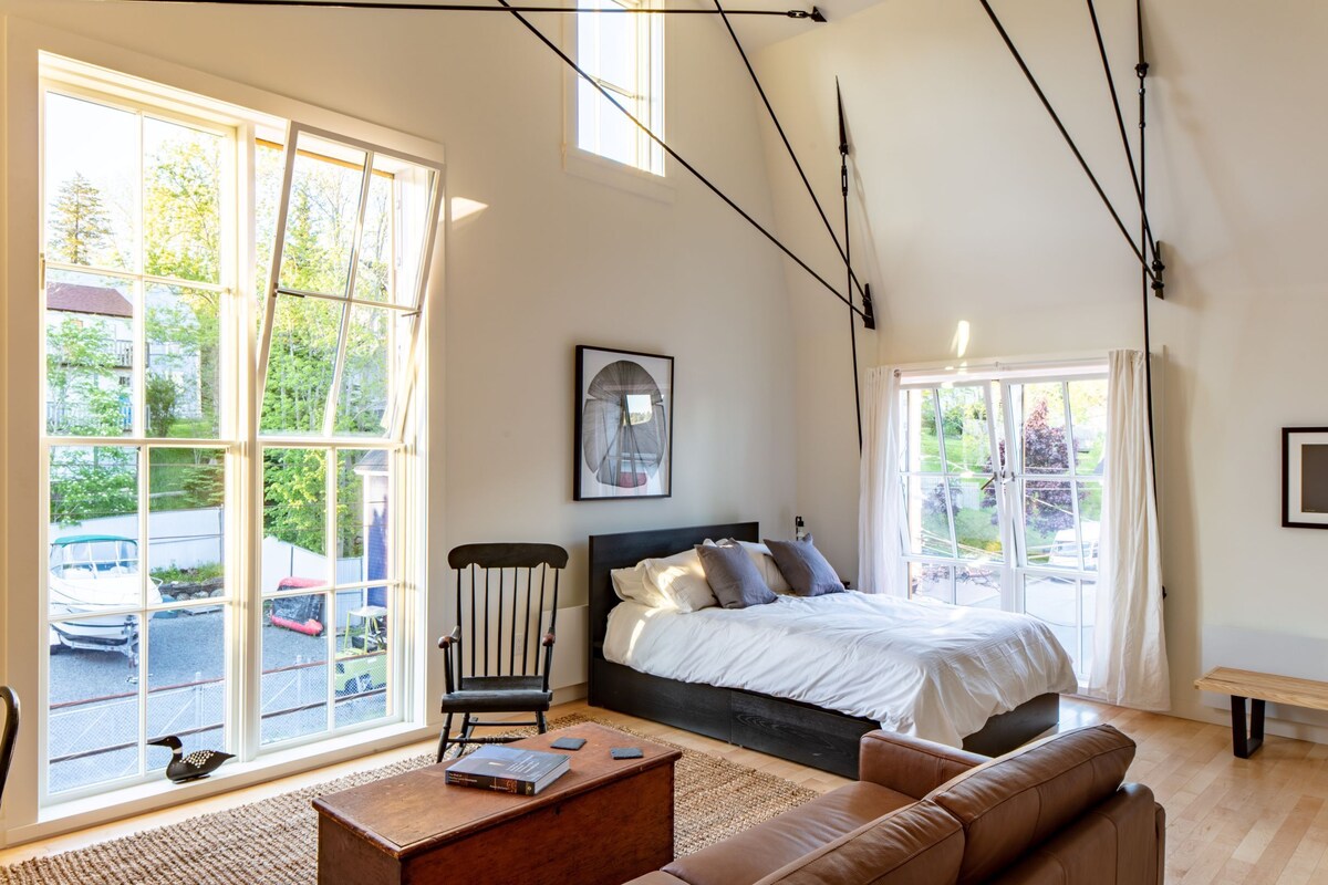 Ein Schlafzimmer mit einem großen Bett, einem Sessel, einer Couch, einem Couchtisch und einem Fenster mit Blick auf parkende Autos und Grünflächen.