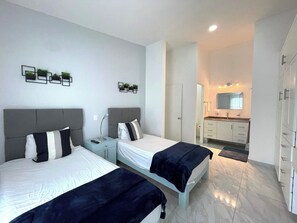 Une chambre d’hôtel moderne avec deux lits, une petite table de chevet avec une lampe, une salle de bain avec un miroir et un lavabo, et une armoire.