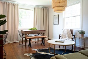 Cozy living room with a round table, bench, and dining table, featuring large windows and a woven light fixture.