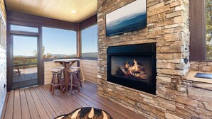 Una chimenea de piedra con un televisor montado arriba, una terraza de madera con una mesa y sillas, y vistas a las montañas a través de amplios ventanales.