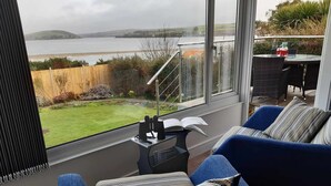 Ett mysigt rum med ett stort fönster med utsikt över en sjö eller ett hav och kullar. Det finns en blå soffa med mönstrade kuddar, ett litet bord med en kikare och en bok, och ett glasräcke.