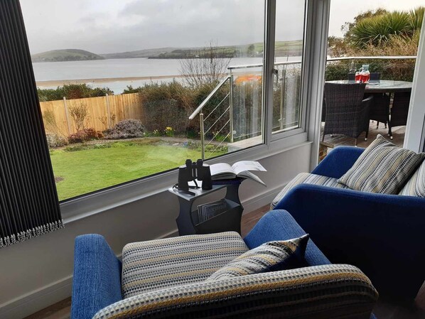 A cozy room with a large window overlooking a body of water and hills. There is a blue sofa with patterned cushions, a small table with binoculars and a book, and a glass railing. - Uninterrupted spectacular views over the Camel Estuary and ocean. (Padstow)
