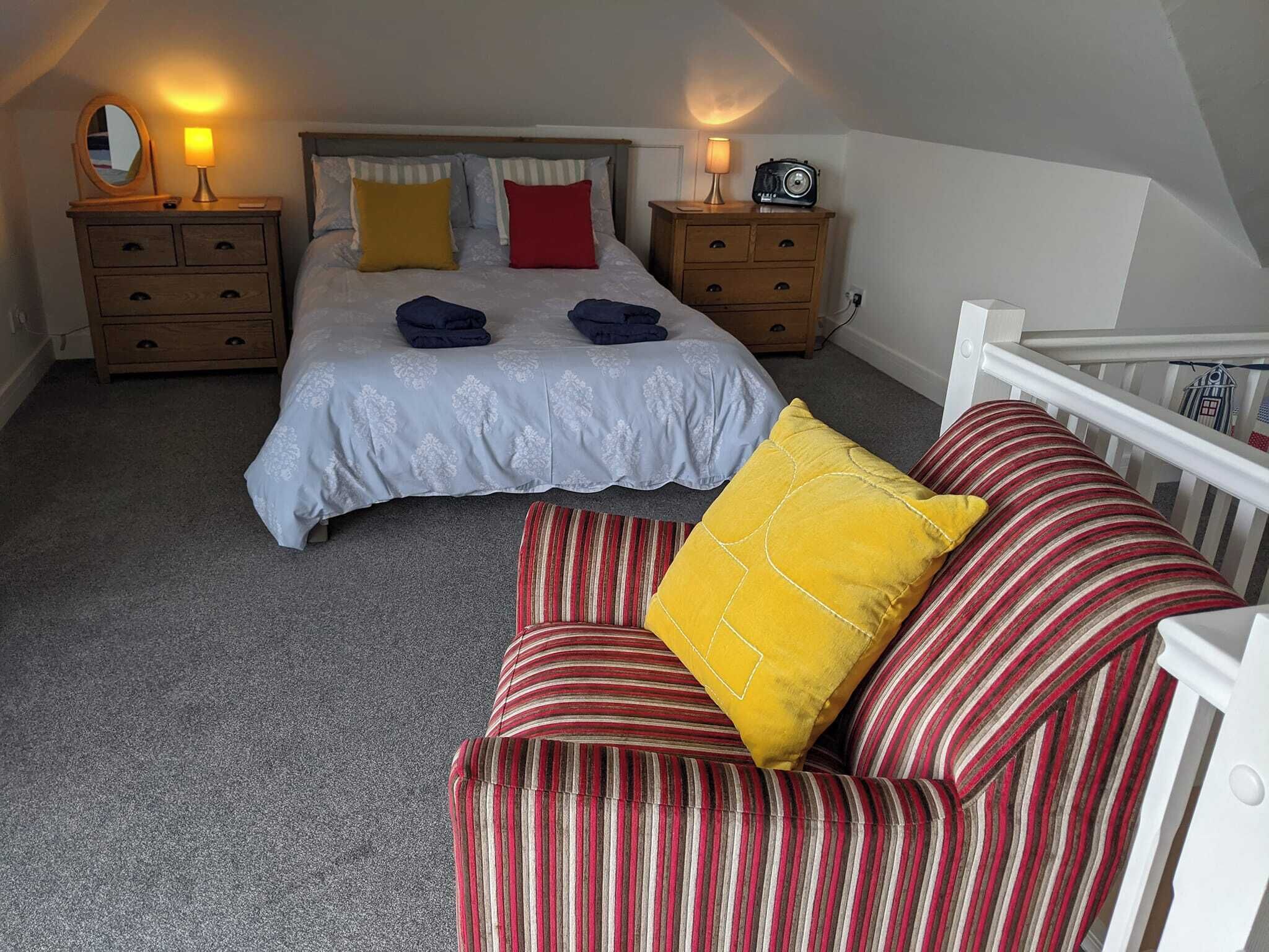 Een slaapkamer met een bed, twee nachtkastjes, een dressoir, een gestreepte stoel en een klein wit kinderbedje.