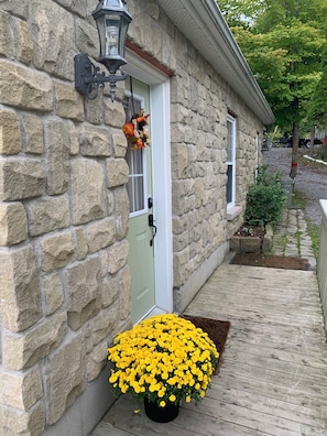 Ein Steinhaus mit einer weißen Tür, einem Laternen über der Tür und einem hölzernen Deck mit gelben Blumen.