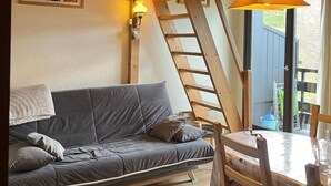 A cozy room with a wooden ceiling, a gray sofa, a wooden dining table with chairs, and a ladder leading to a window.
