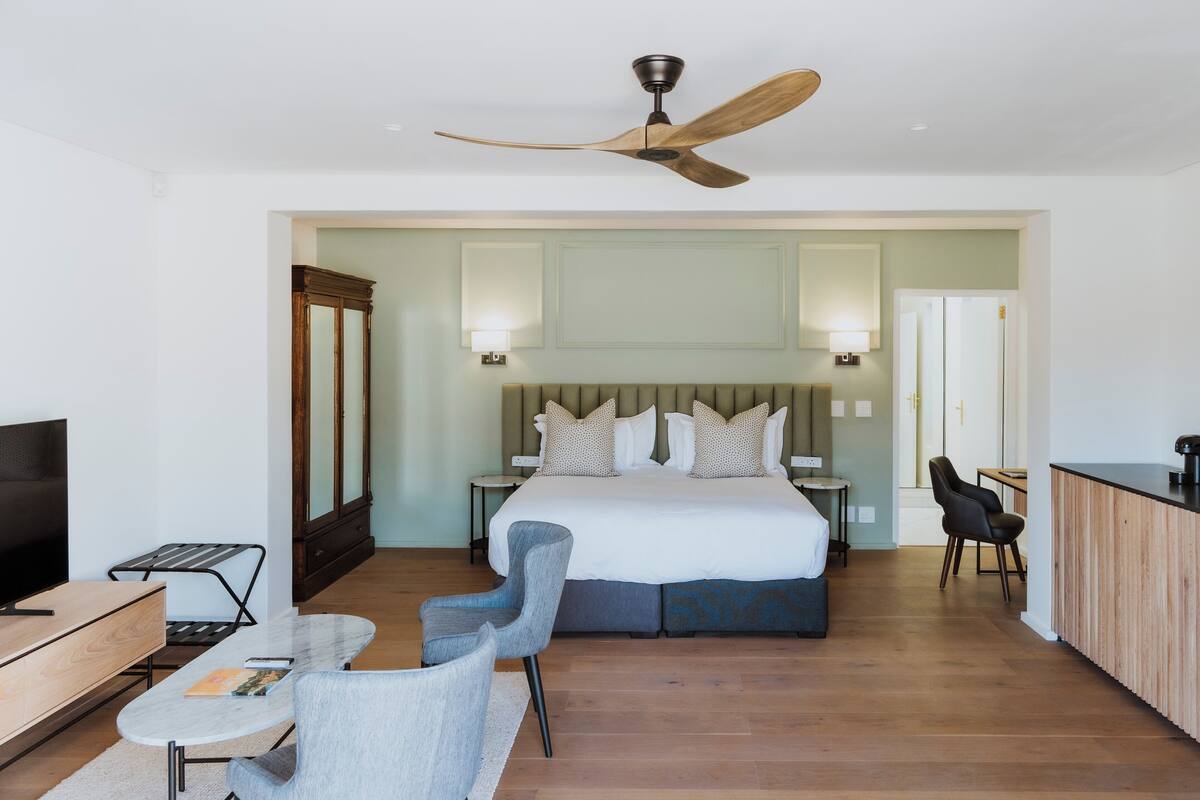 A modern hotel room with a large bed, a desk, a chair, a TV, and a ceiling fan.