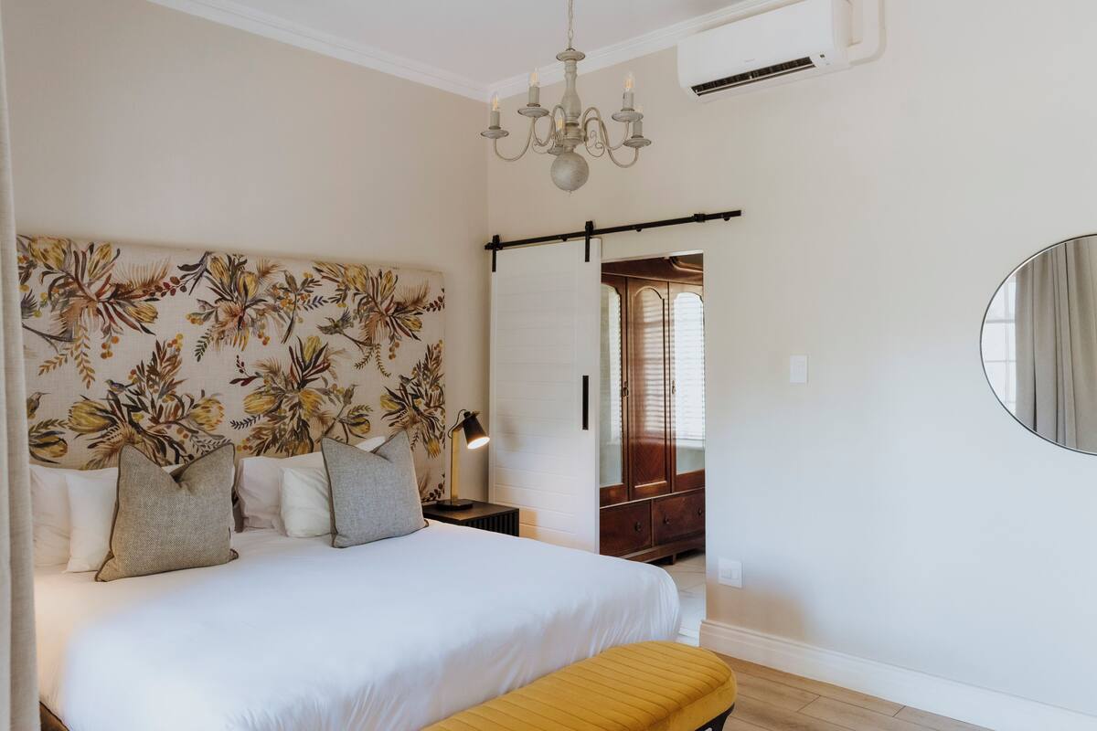 A bedroom with a large bed, a headboard with a floral pattern, a bedside table with a lamp, a mirror on the wall, and a wooden bench.