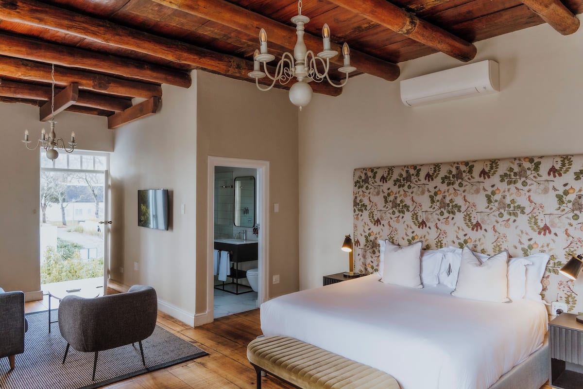 A bedroom with a large bed, a chandelier, a bedside table with a lamp, and a view of the bathroom.