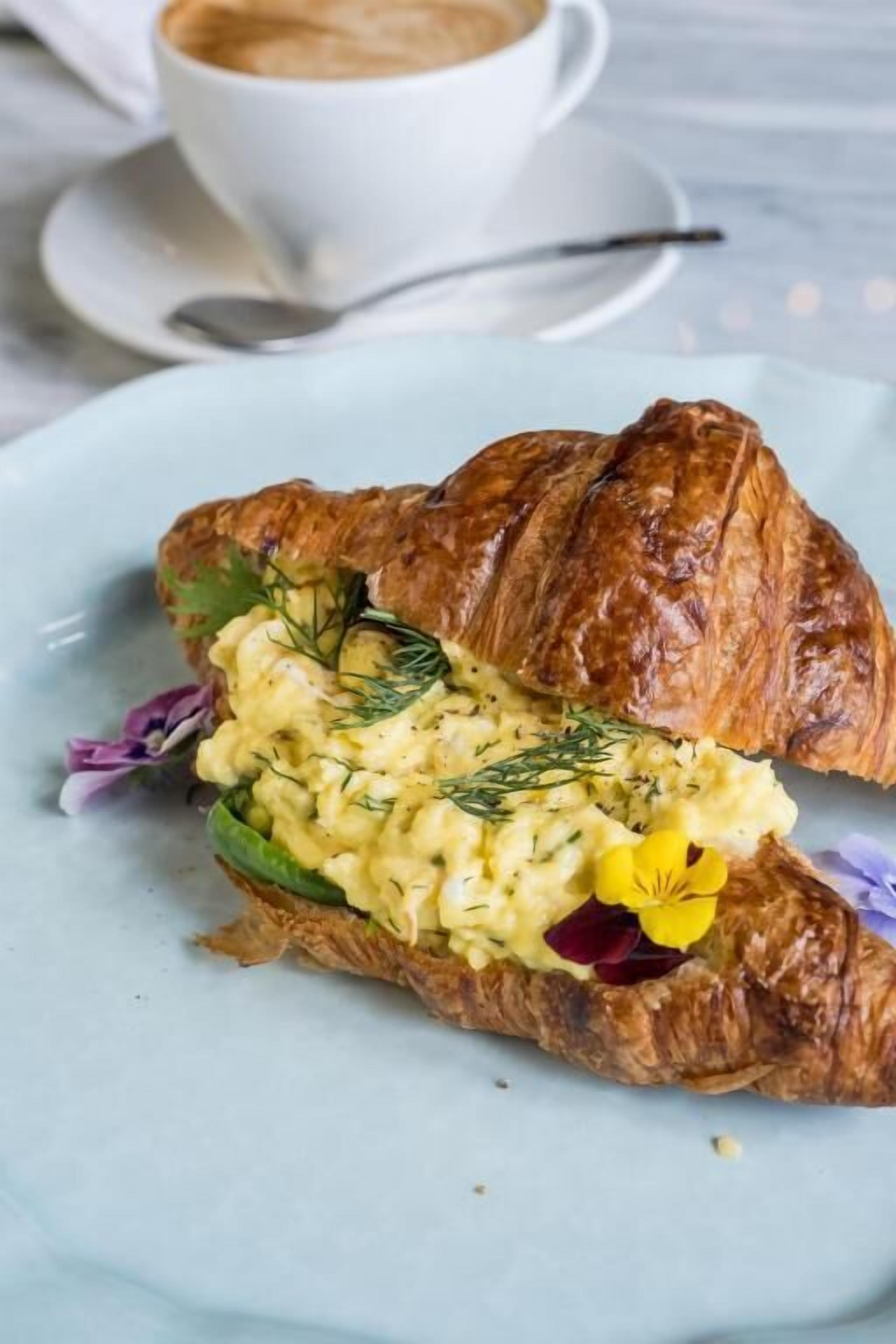Ein Croissant-Sandwich mit cremiger Füllung und frischen Kräutern, serviert auf einem hellblauen Teller.
