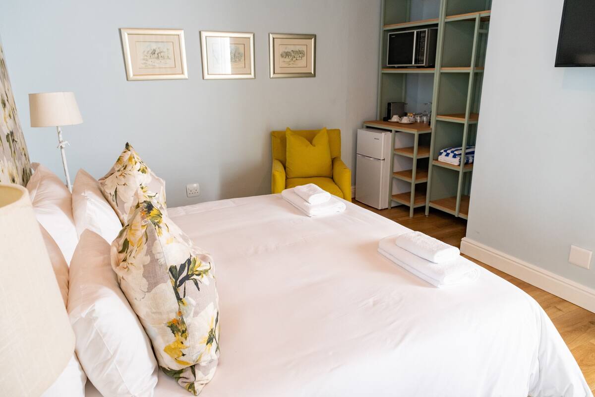 A neatly made bed with white linens, a yellow armchair, a small white refrigerator, and a shelving unit with various items.