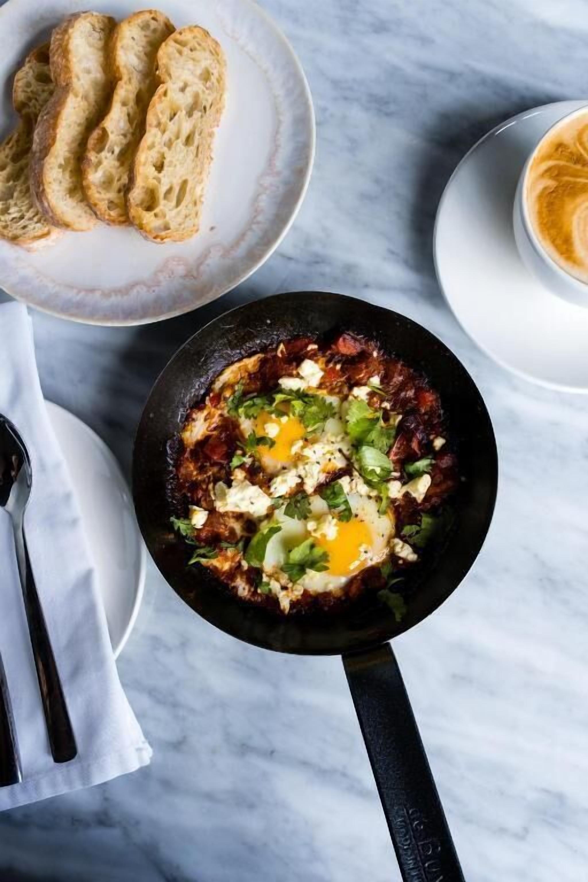 A breakfast setting with a skillet of eggs and vegetables, a plate of bread, and a cup of coffee on a marble surface.