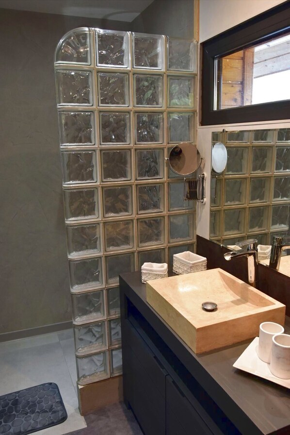 Un baño moderno con cabina de ducha de vidrio, tocador de madera con lavabo color beige y un espejo grande sobre el lavabo.