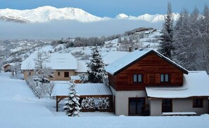 A wooden chalet with a snow-covered roof, surrounded by a snowy landscape and mountains in the background.