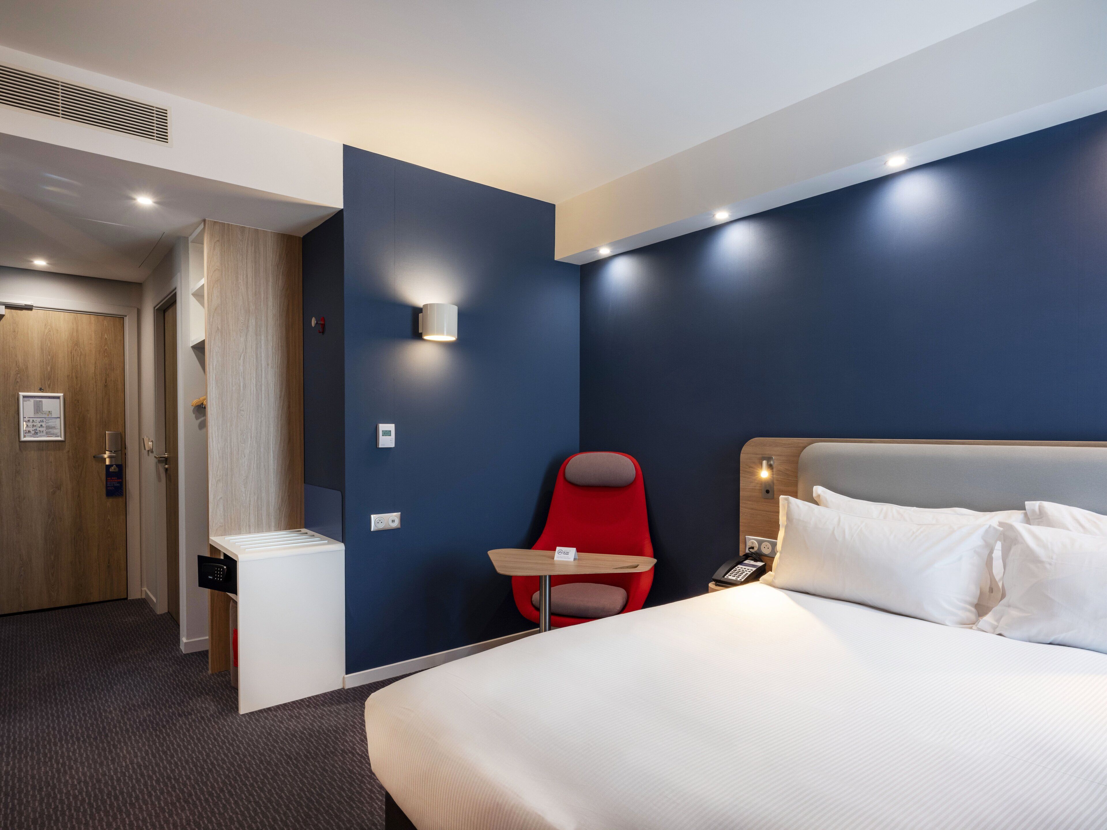 A hotel room with a bed, a red chair, a small table, and a wall-mounted phone.