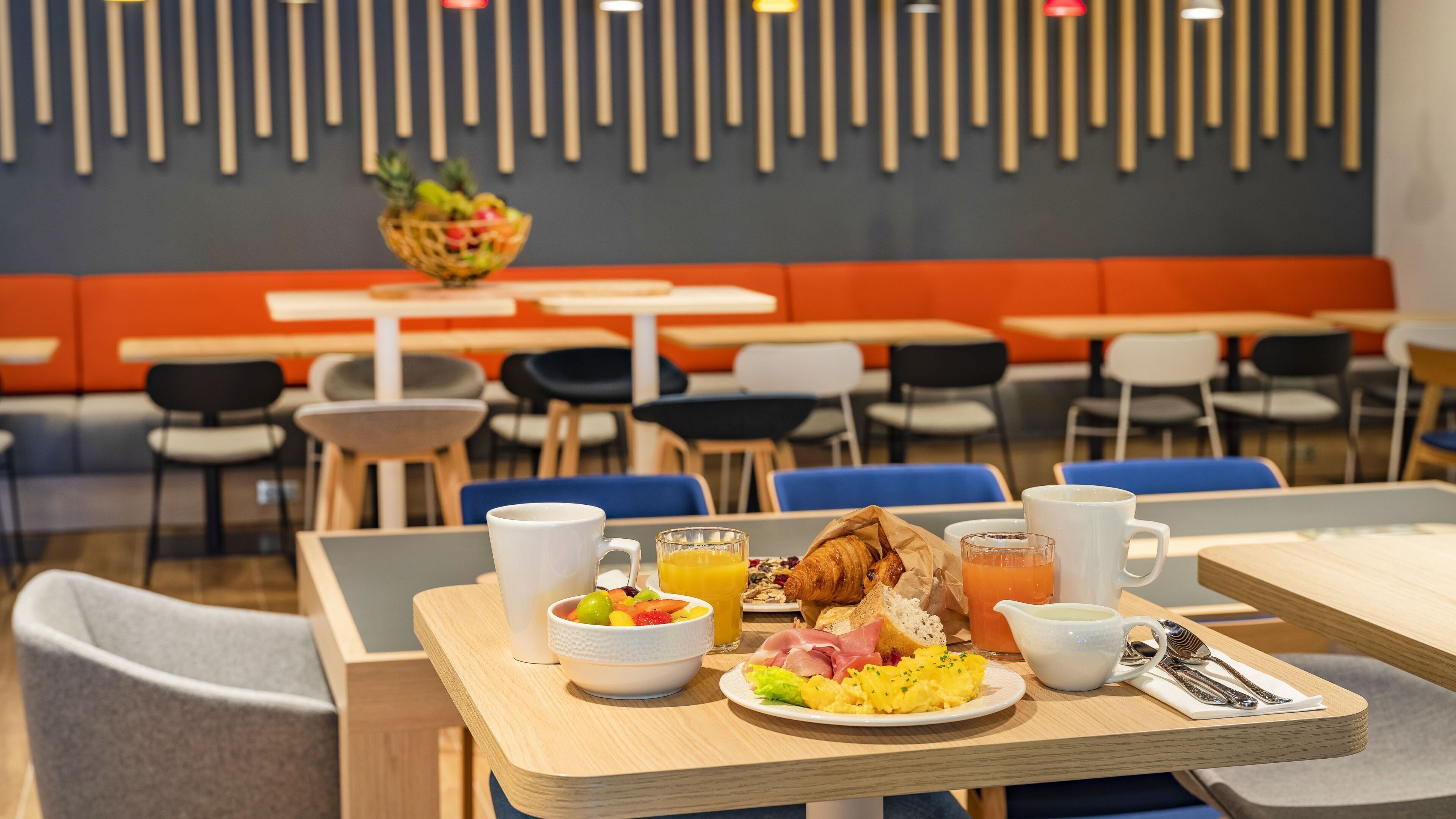 Une table mise pour le petit-déjeuner, avec une place pour deux personnes, comprenant un croissant, un assortiment de fruits, du jus d’orange et des œufs brouillés.
