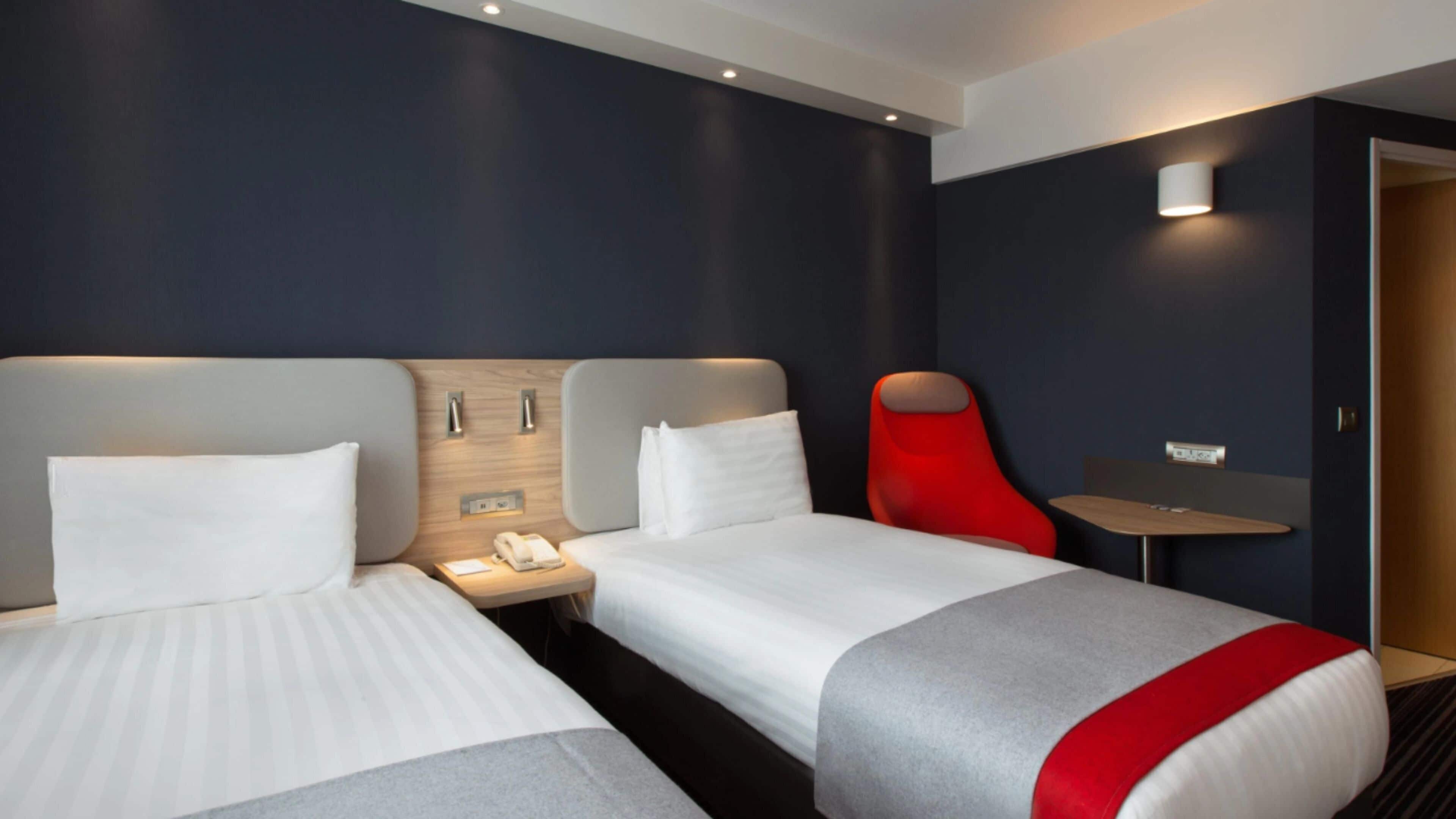 A hotel room with two beds, a red chair, a small table, and a wall-mounted phone.