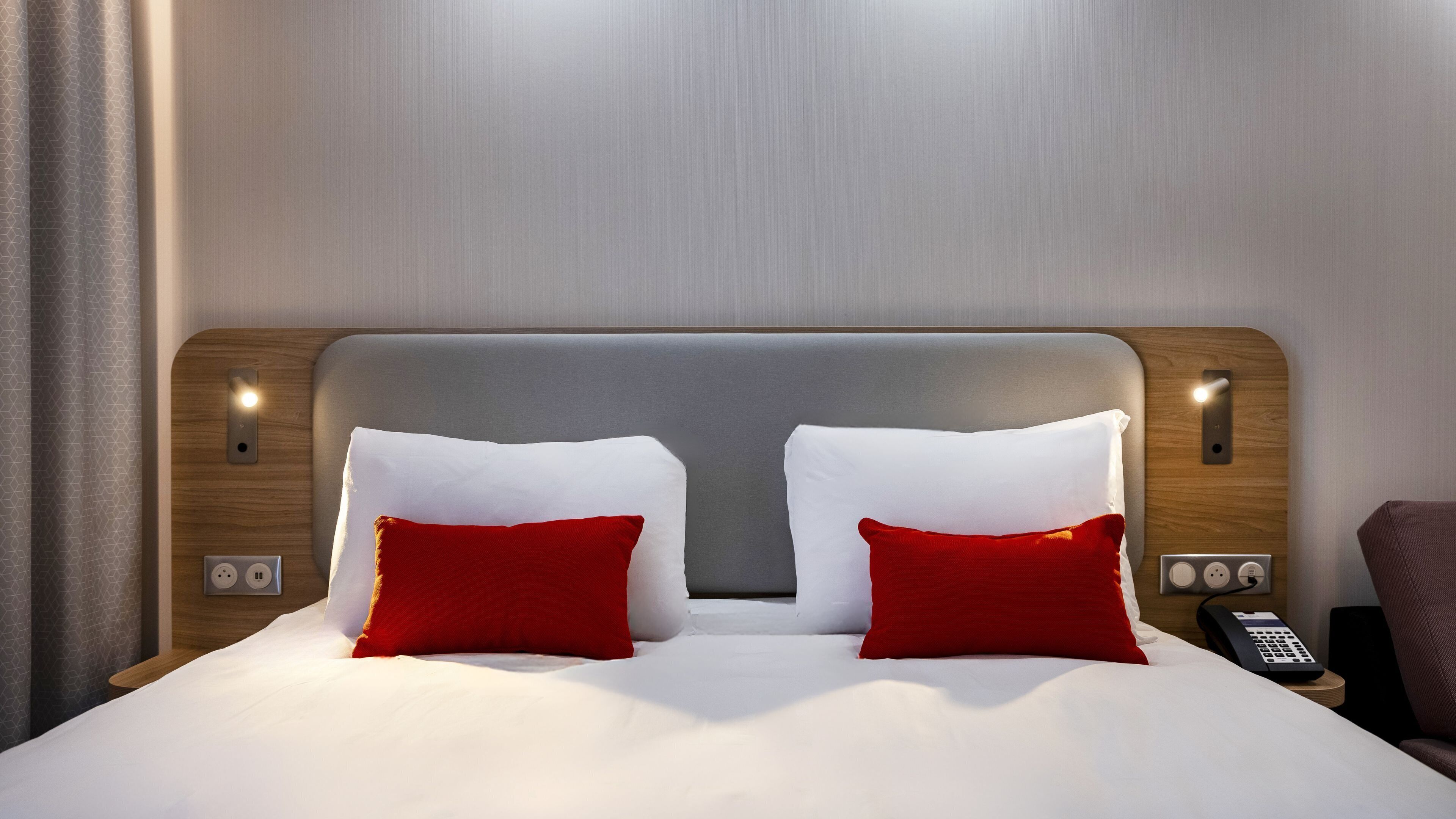 Een hotelkamer met een groot bed, twee rode kussens, een houten hoofdbord, nachtlampjes en een telefoon.