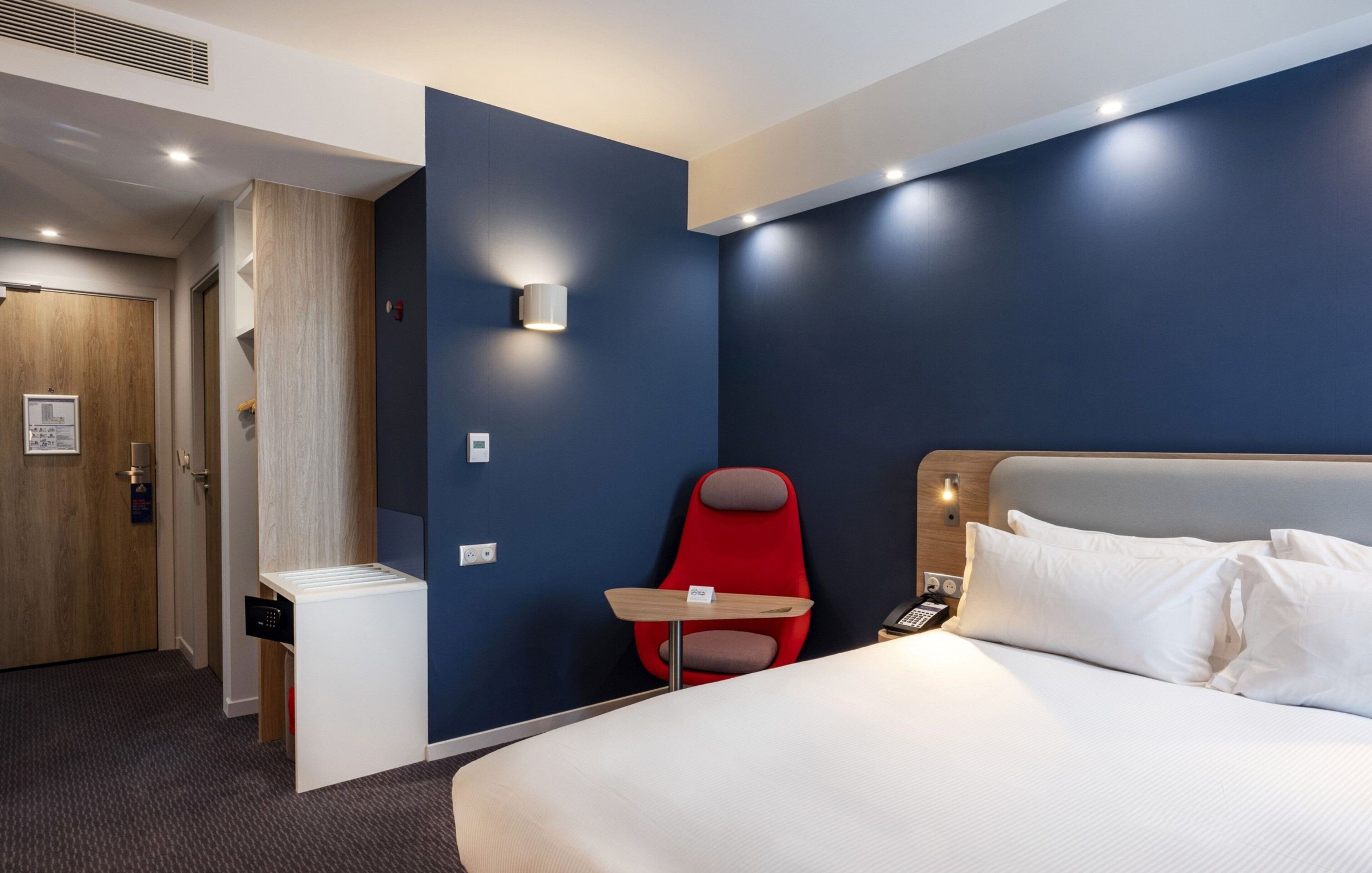 Une chambre d’hôtel avec un lit, une chaise rouge, une petite table et un téléphone fixé au mur.