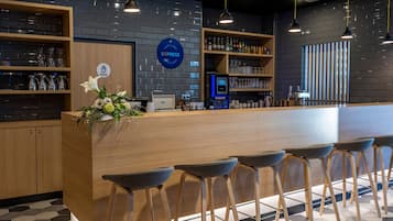 Zona de bar moderna con mostrador de madera, taburetes y una exhibición de botellas y vasos.