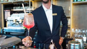 A bartender holding a cocktail with a straw, behind him is a bar with various bottles and glasses.